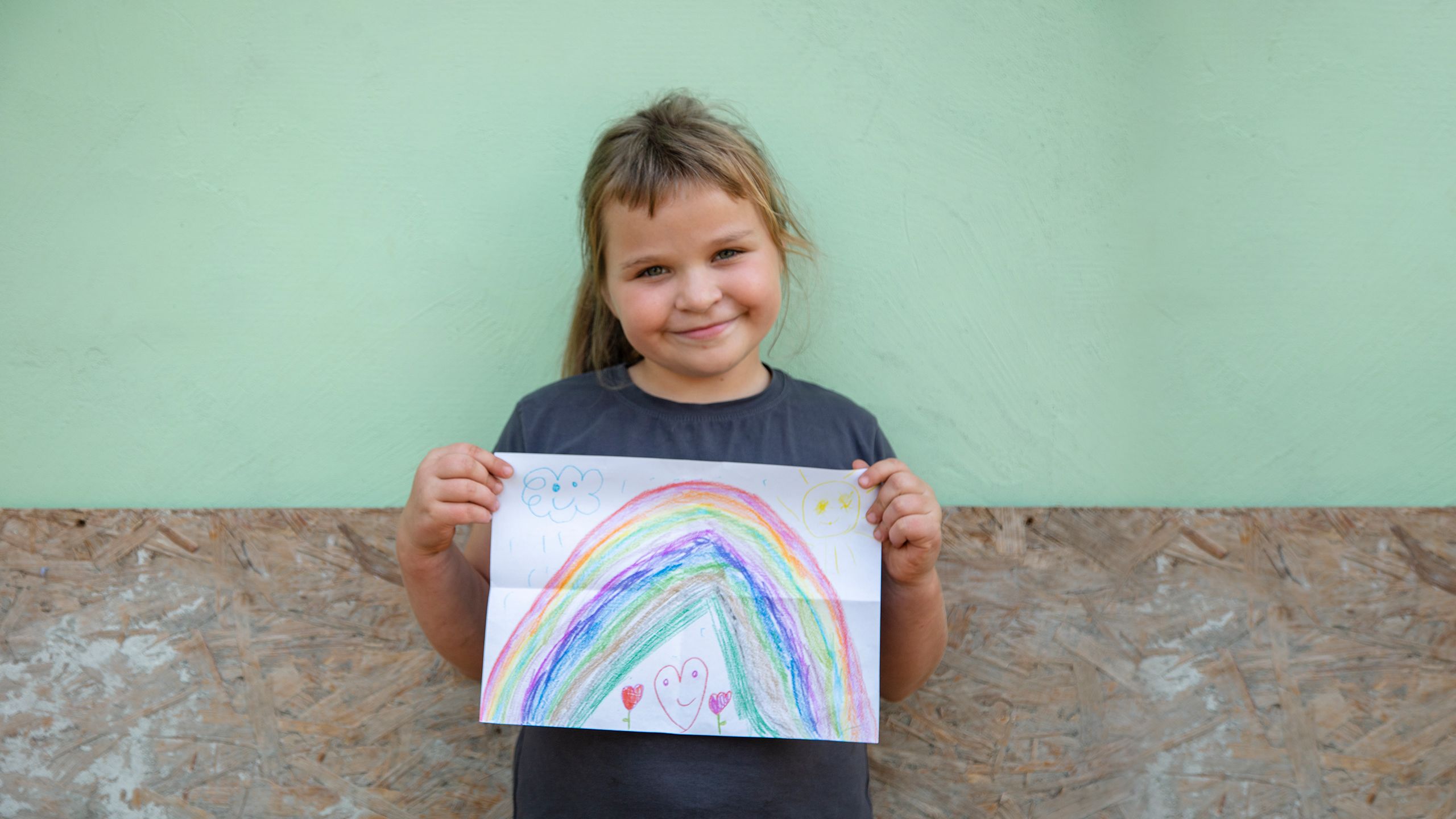 A young girl smiles and holds up her drawing of a rainbow in front of her house.