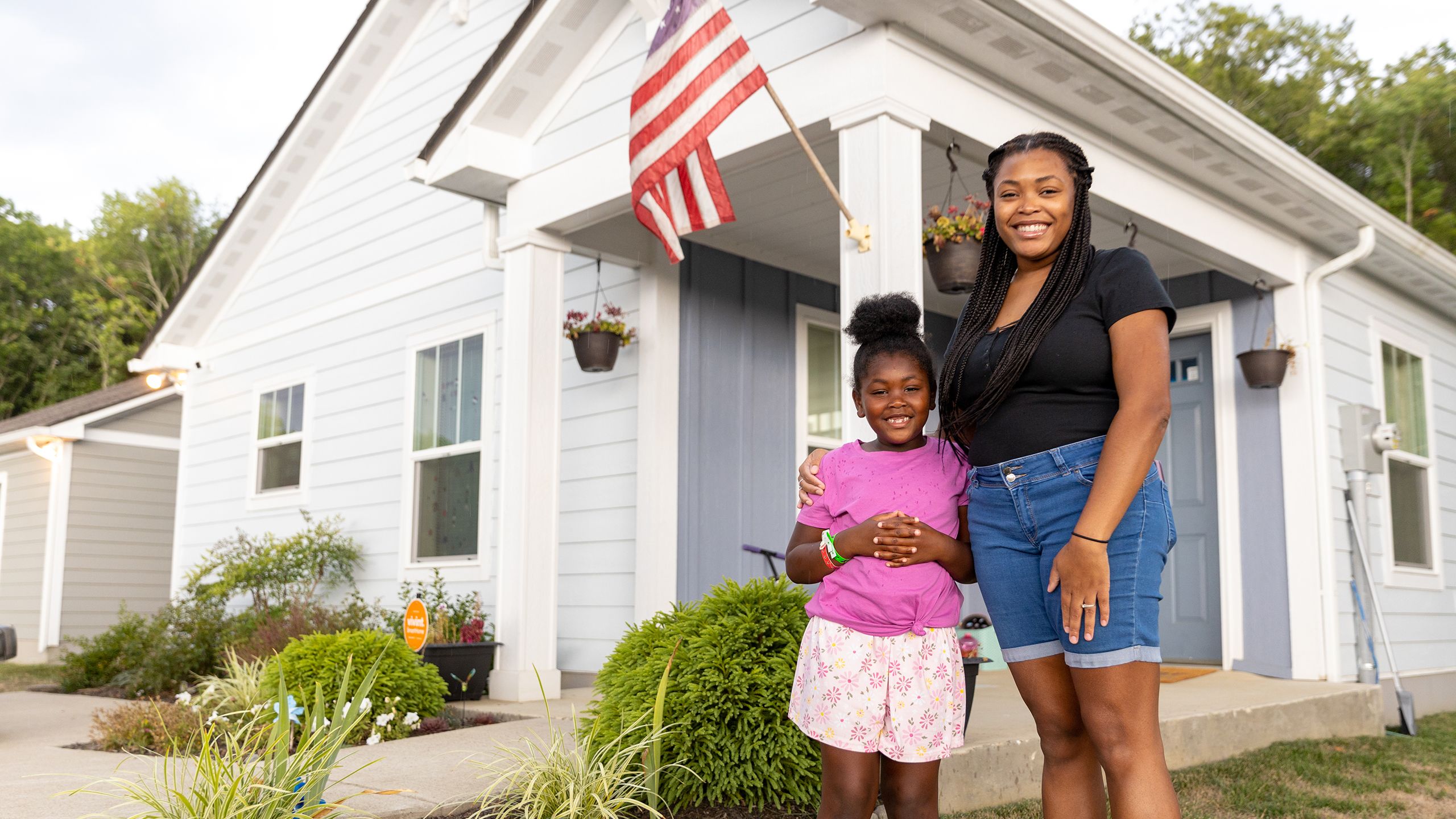 D'Shawna, a young Black woman, stands proudly with Ariana in the thriving front garden of their pale gray Habitat house.
