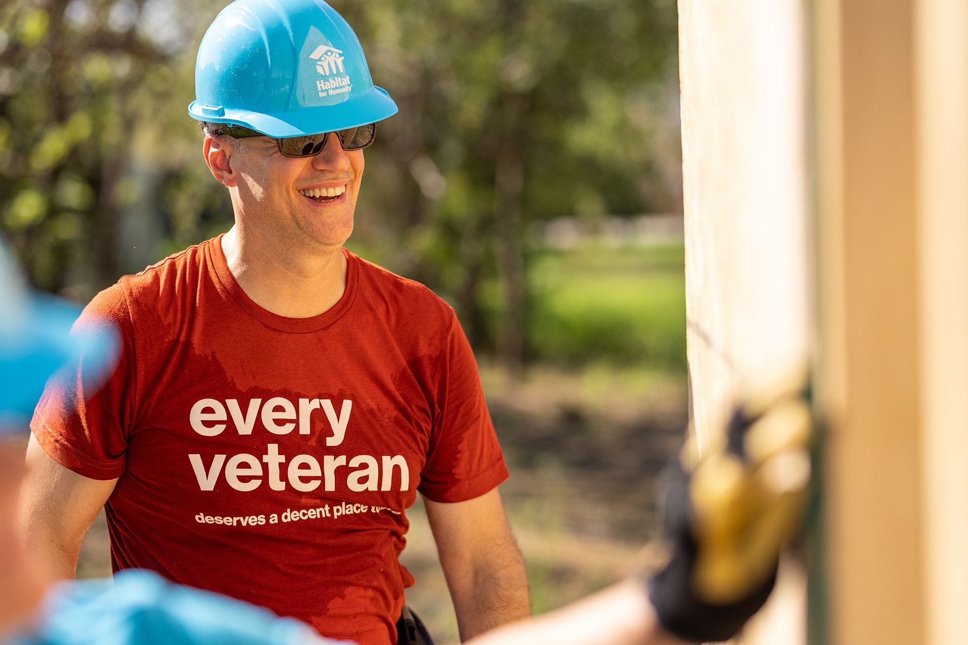 A smiling volunteer wearing a t-shirt reading "every veteran deserves a decent place to live" helps attach plywood to a wall