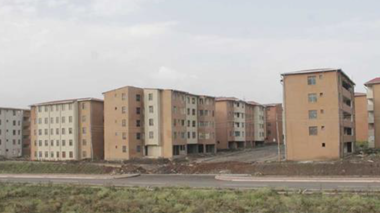Four apartment-style buildings with beige outer walls and four floors of windows and balconies and a sidewalk in front of it.