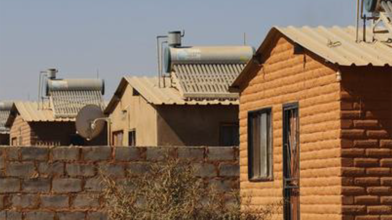 One brown house in the foreground alongside a brick wall with two rooftops visible behind it against a blue sky.