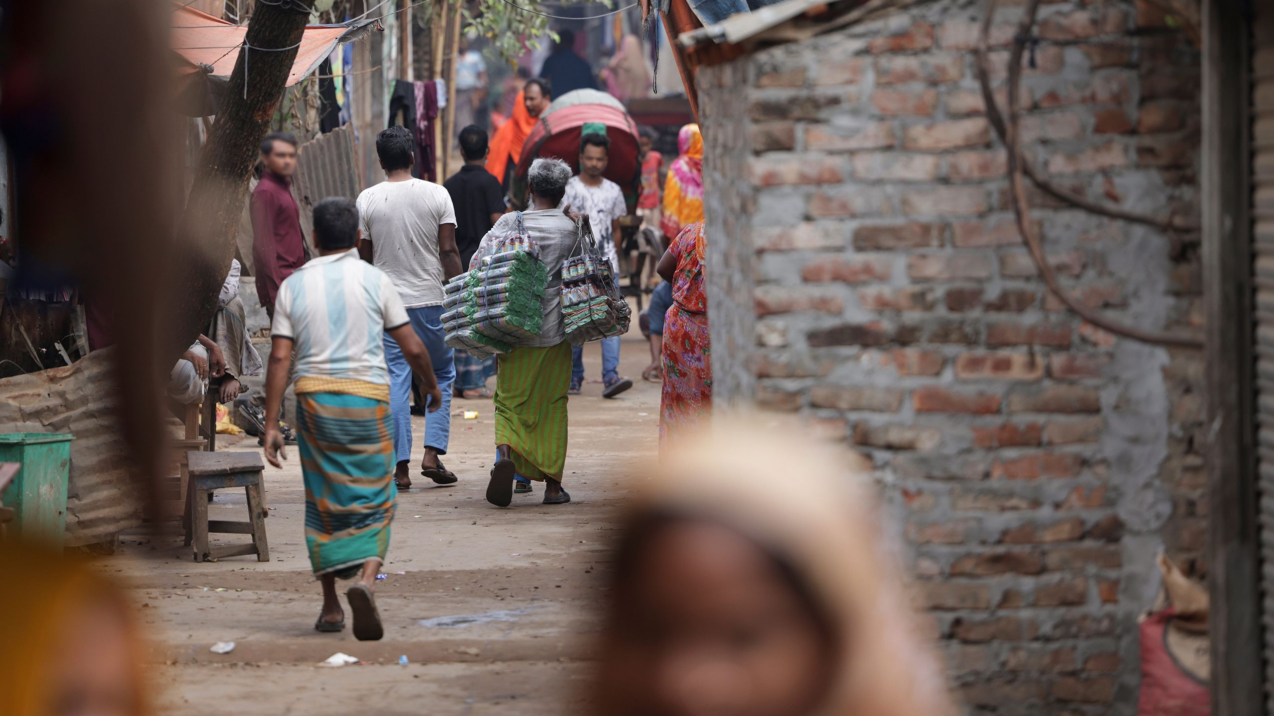 Seen from behind, a crowd of people walks down a passageway in Beguntila, carrying items, pulling carts and doing other daily tasks.