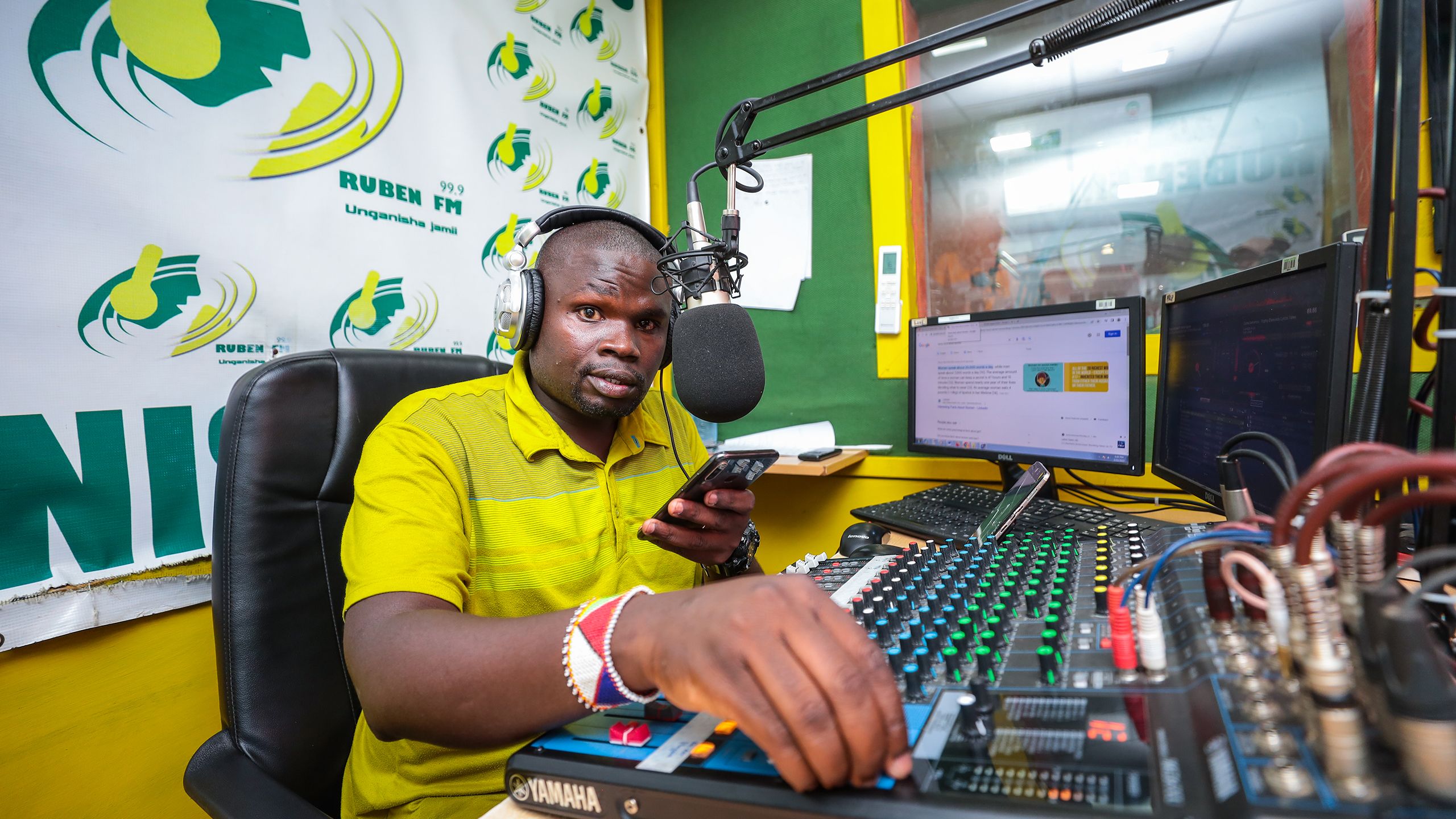 Robert, a young Kenyan man in a bright yellow polo shirt, is at work in his radio studio. He talks into a cellphone held in one hand near his microphone as he adjusts levels on his soundboard with his other hand..