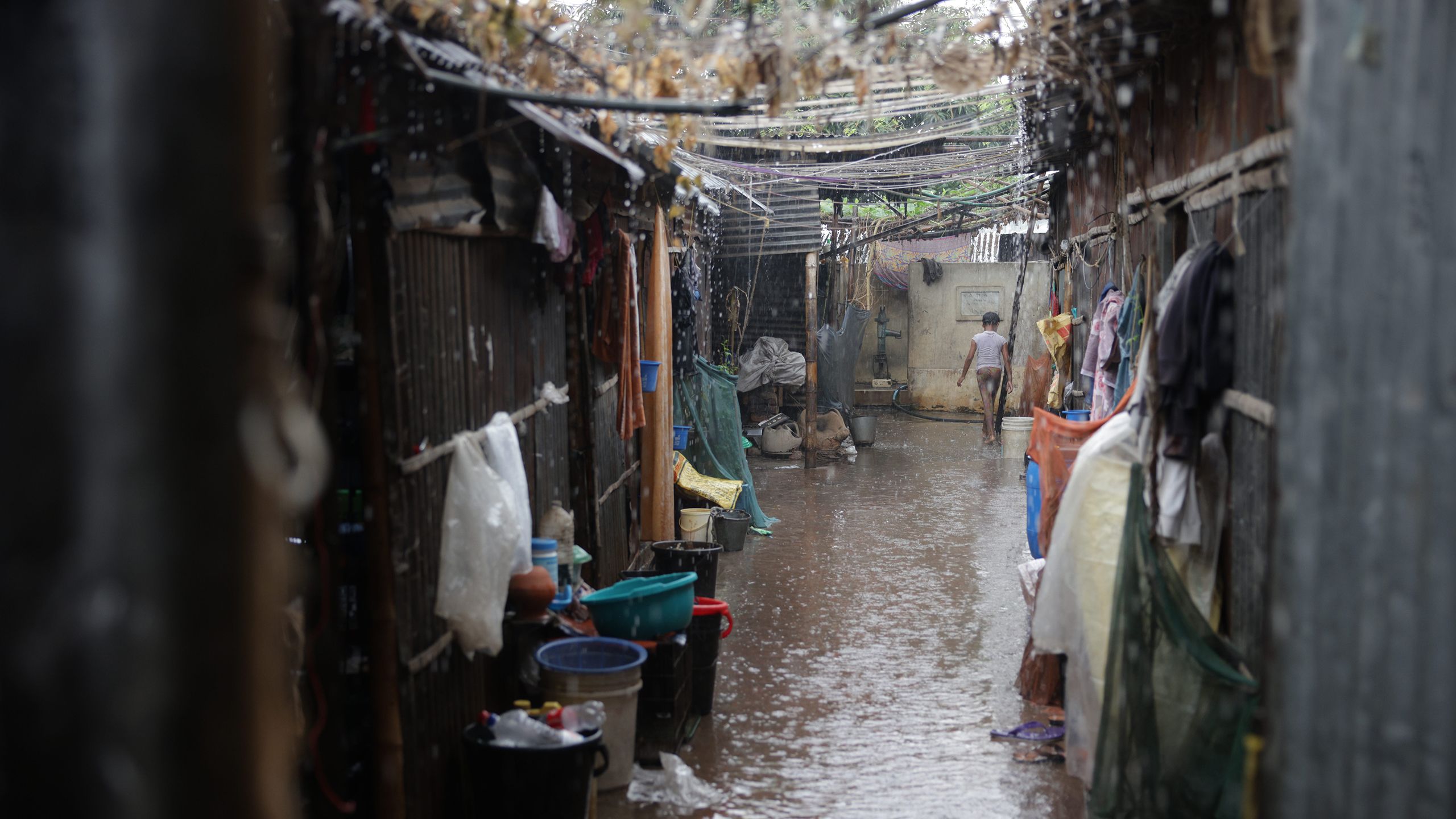 Rain floods the narrow path between small homes. At the far end of the path, a young boy walks in ankle-deep water.