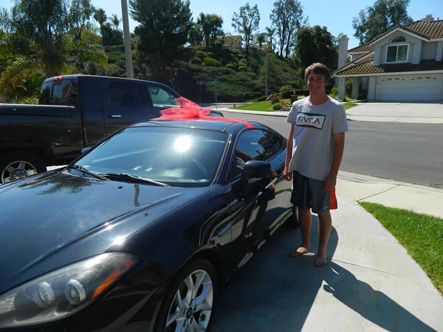 Black Hyundai Tiburon with a red bow on top. A male teenager stands to the right of the car.