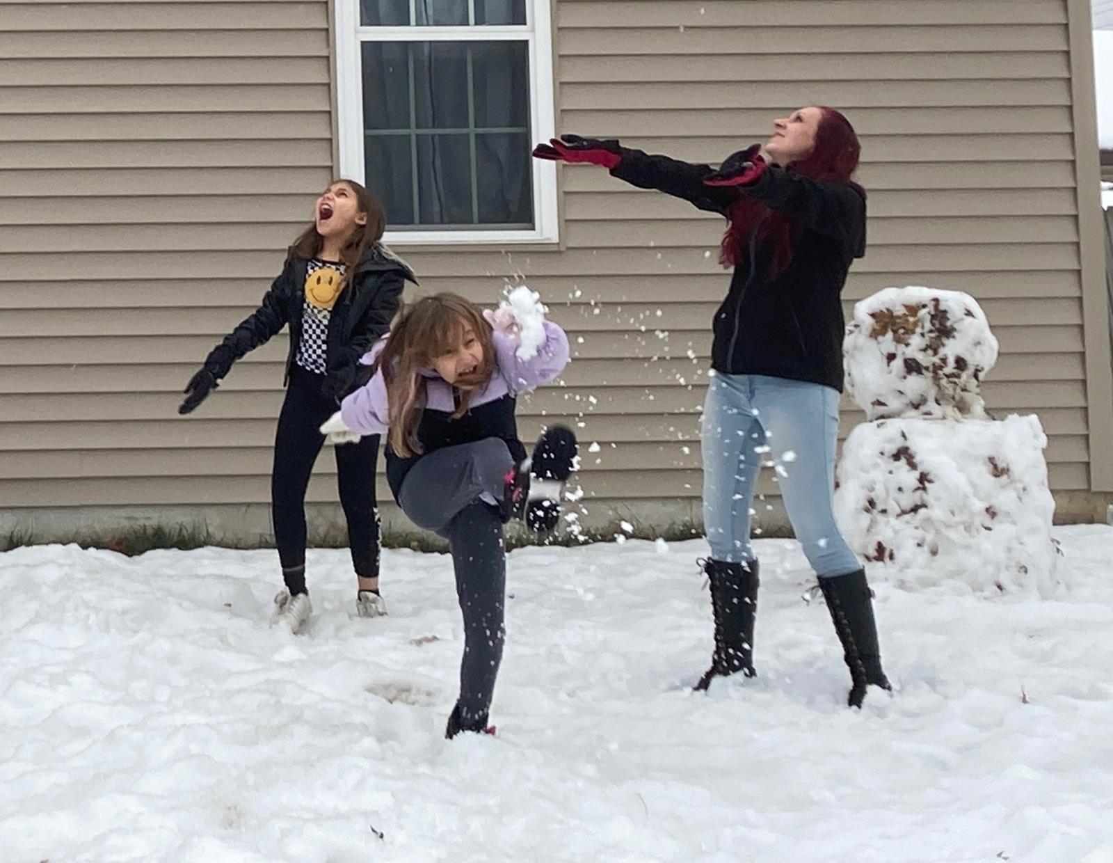 Two young daughters in jeans, boots, and coats playing in the snow next to a snowman with their mom. 