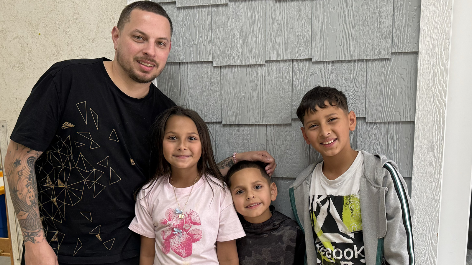 A father and his three kids stand in front of their home and smile at the camera.