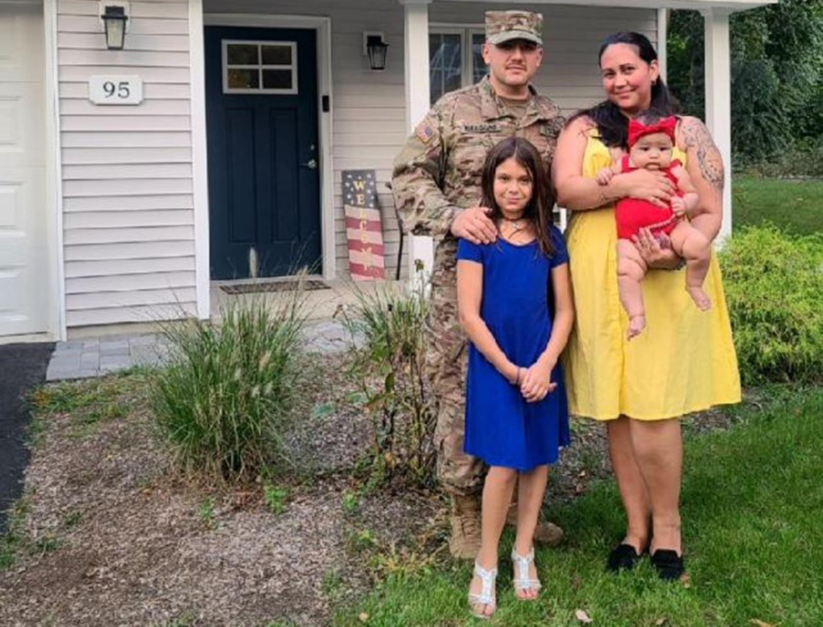 Man in a military uniform posing with his wife in a yellow dress, daughter in a blue dress and infant daughter in a red outfit in front of their home.