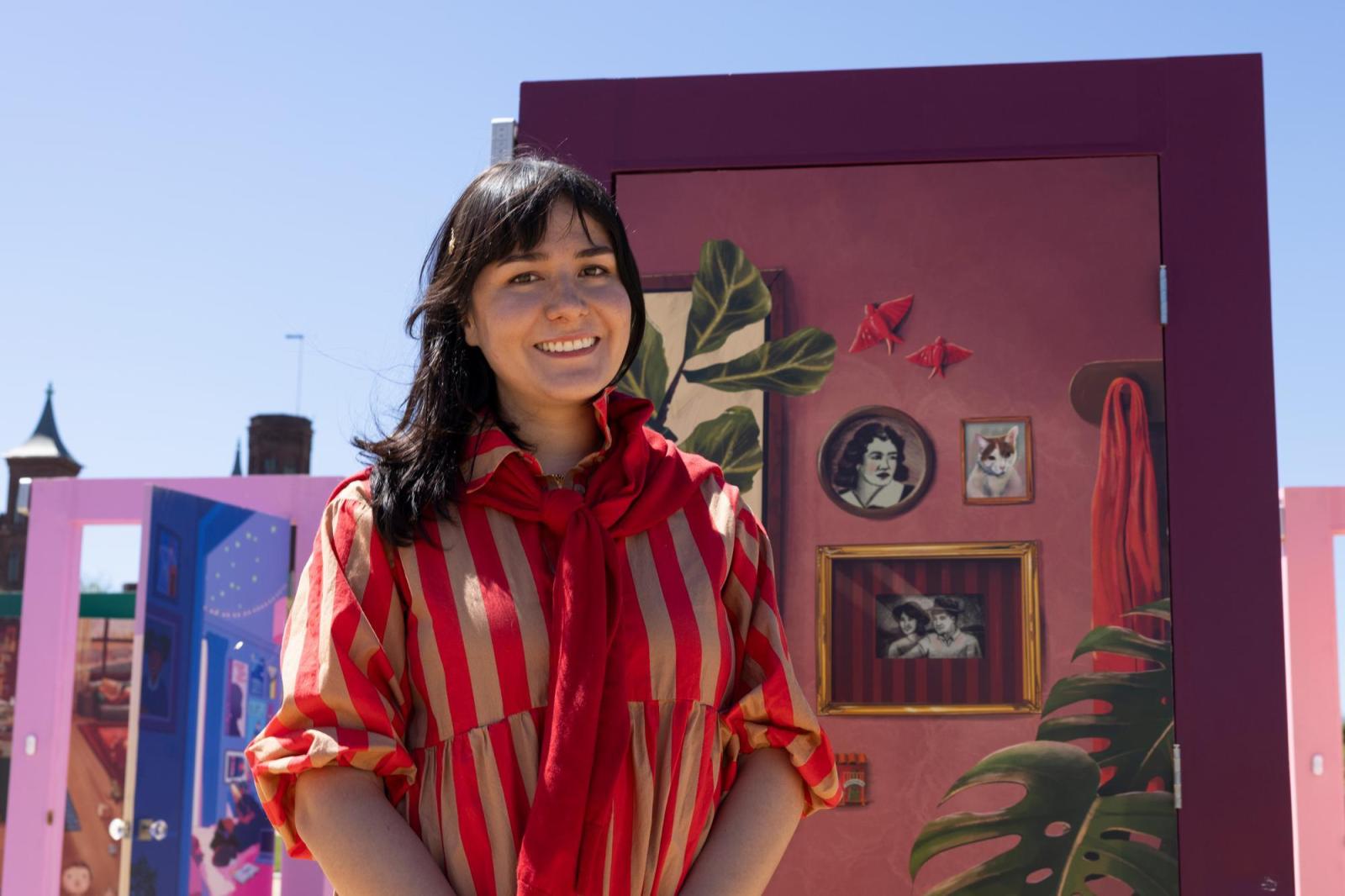 Woman in a red and yellow swiped dress with brown hair standing in front of a pink and purple door with an illustration of photo frames on it.