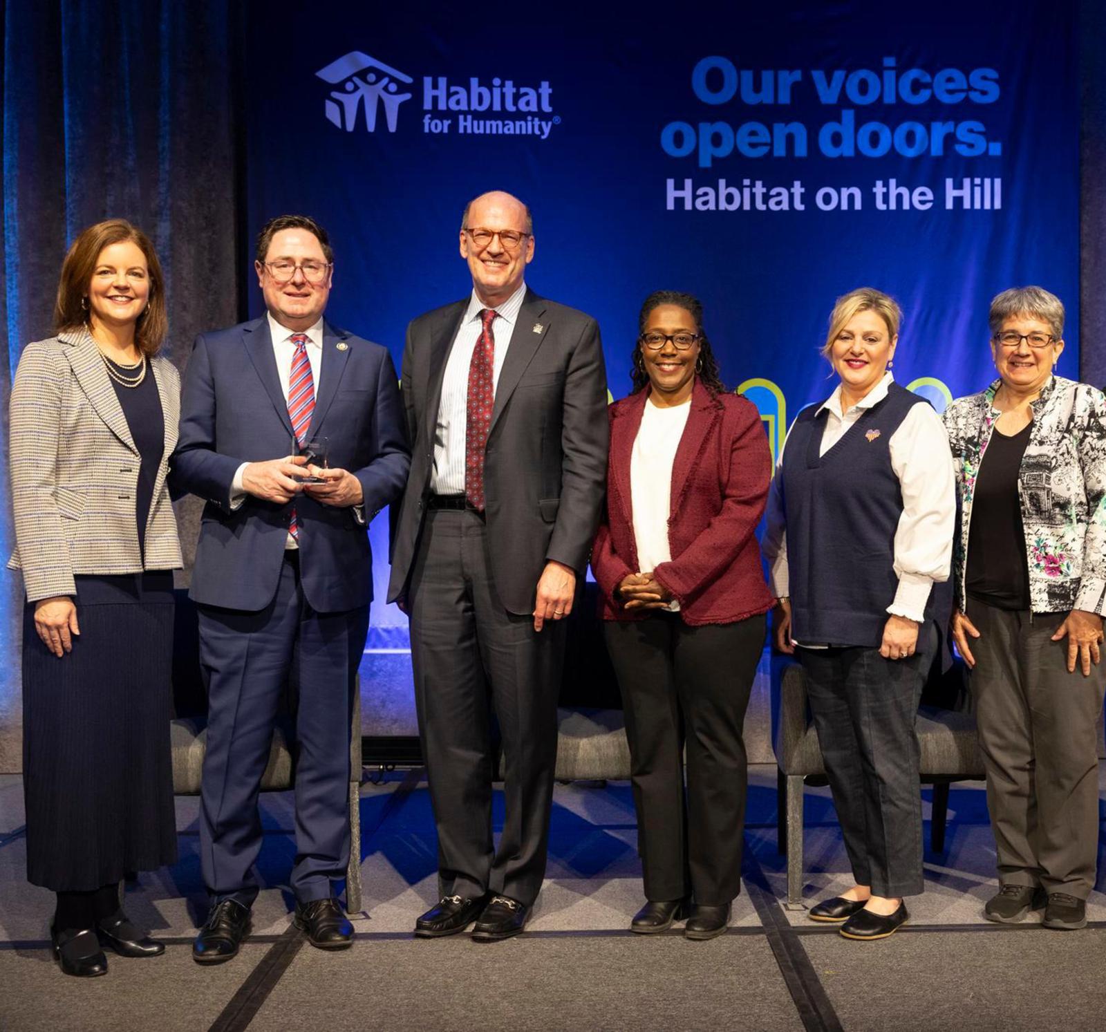 A group of people posing on stage with "our voices open doors, Habitat on the Hill" on a screen in the background.