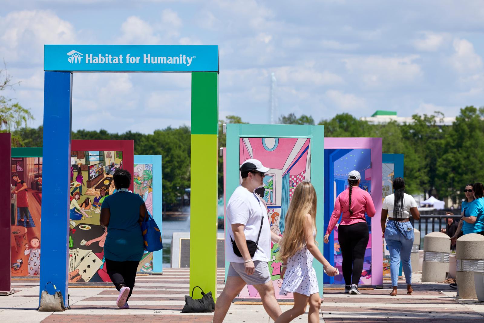 A man and a woman walk by the pop-up door exhibition while another woman walks through a framed opening that says Habitat for Humanity at the top.