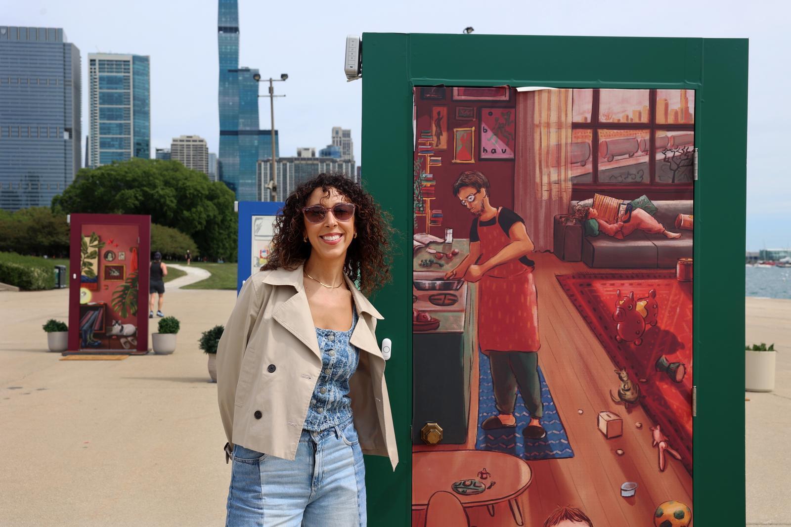 The artist, a woman in a tan jacket and jeans posing next to an illustrated door with the green frame.