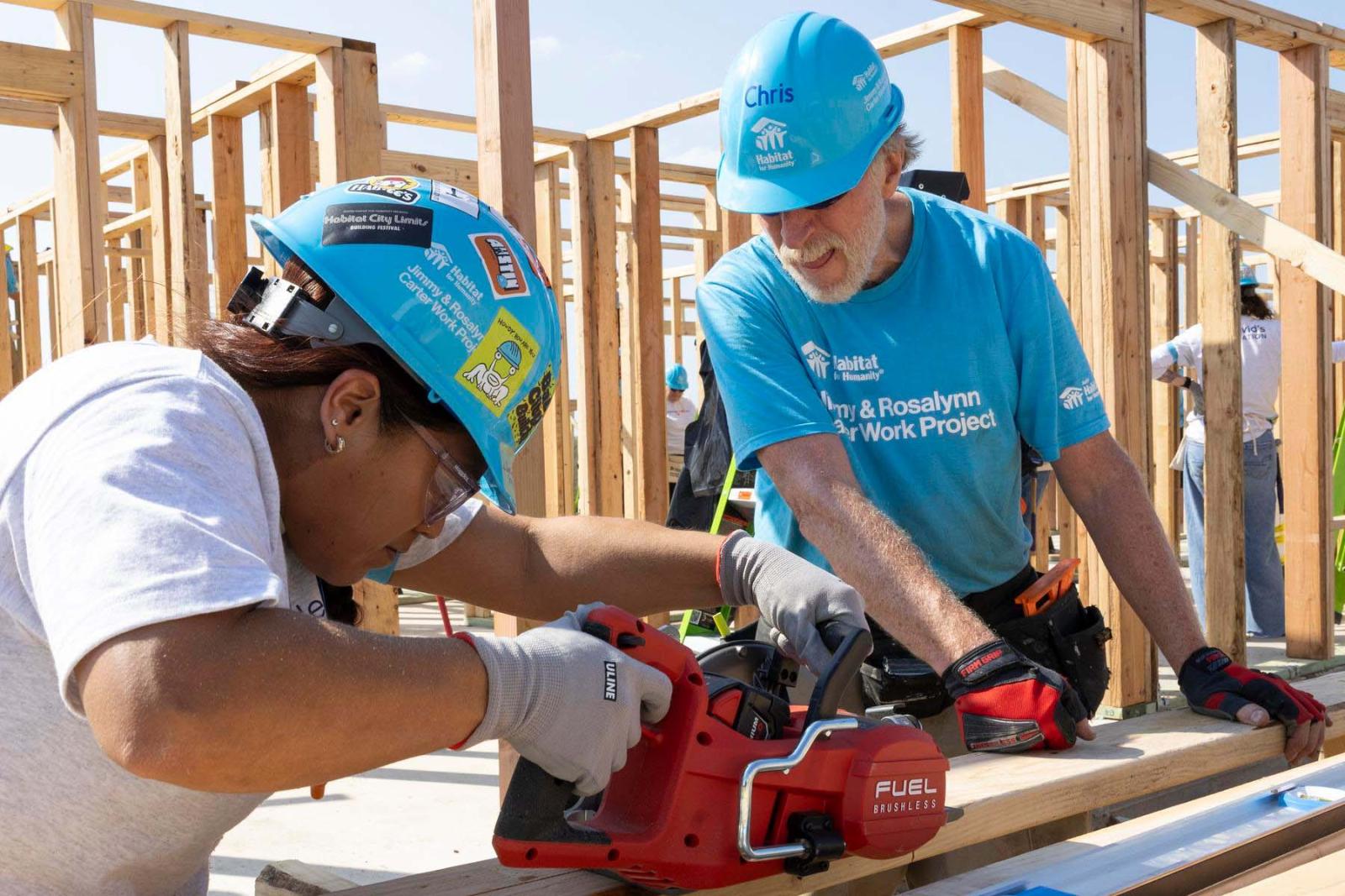 One volunteer stands and uses a power saw on a piece of lumber while the other volunteer holds it in place.