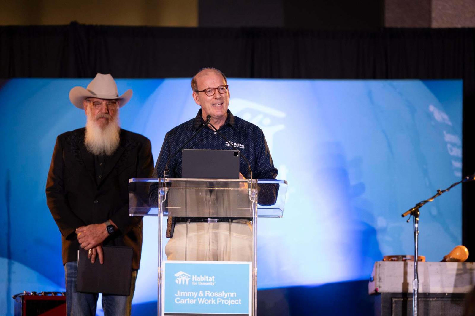 Jonathan Reckford, CEO, Habitat for Humanity International stands alongside Ray Benson, Austin musician and event emcee, behind a podium and microphone on a stage.