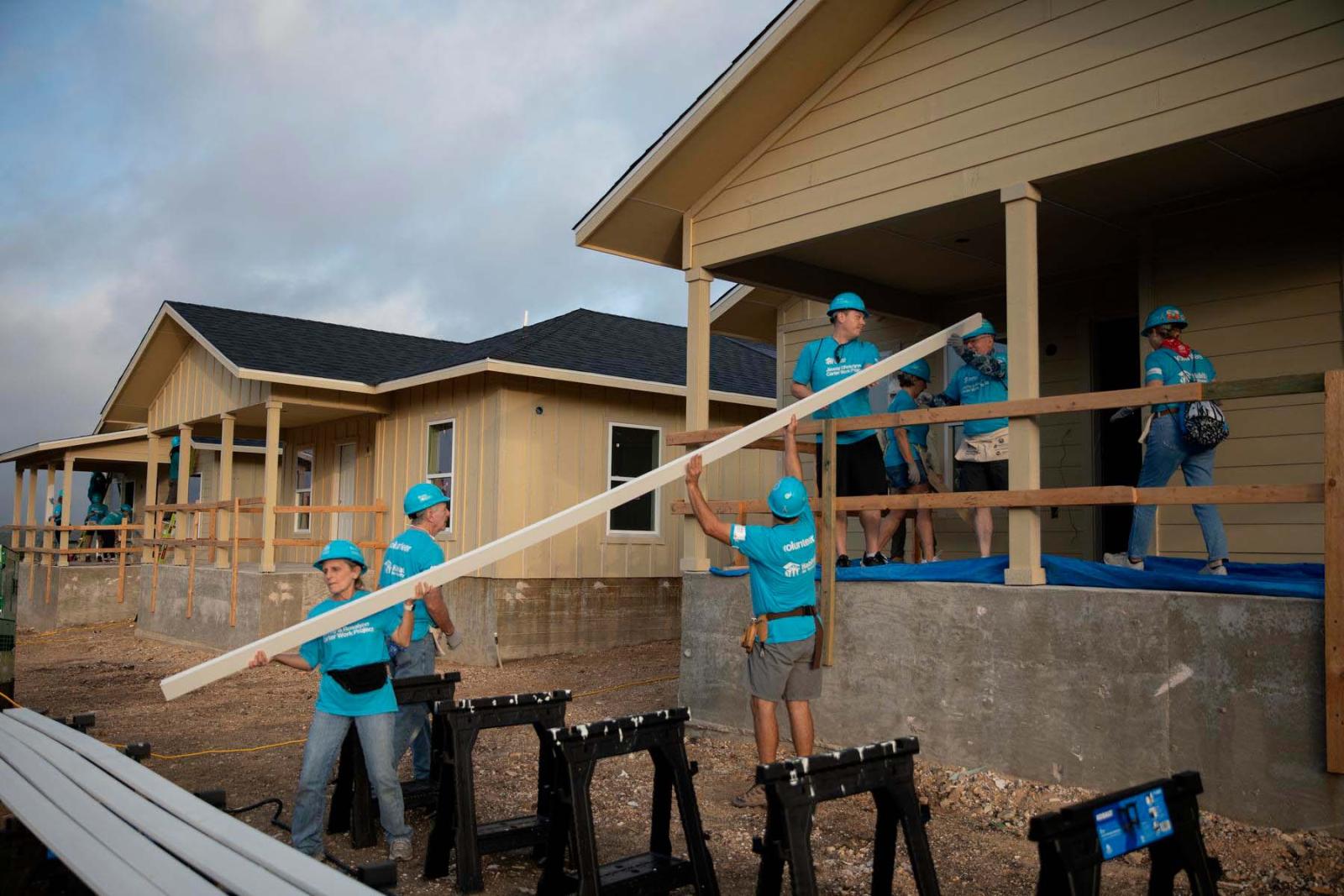 Three volunteers stand on the ground and hold a piece of lumber that is also being held on the other end on the porch of a build house.