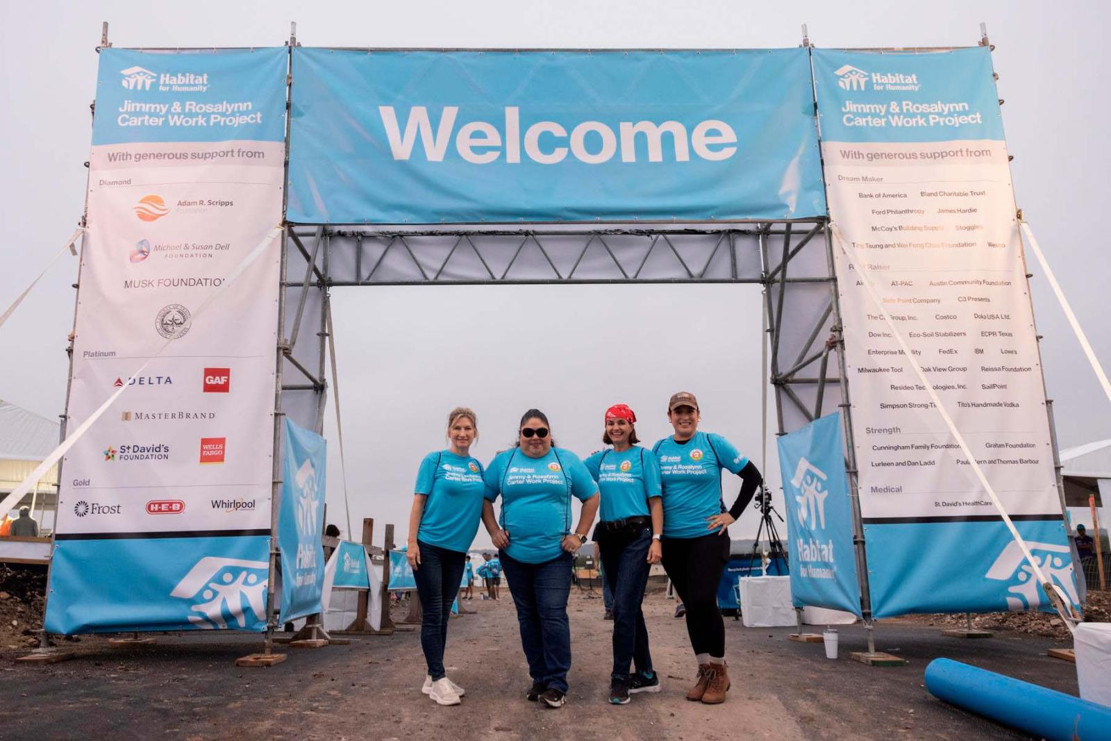 Four volunteers pose in blue Habitat shirts under a giant welcome sign banner.