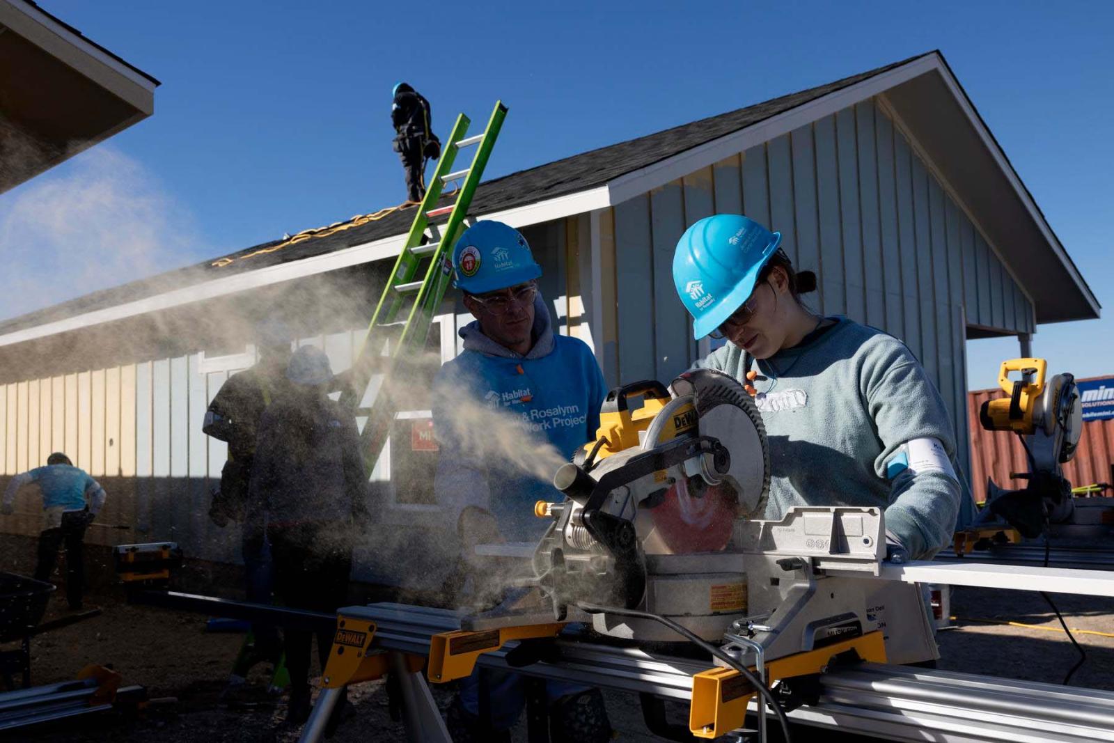 One volunteer in a hard hat uses a power saw and dust floats away as another volunteer looks on in the background.