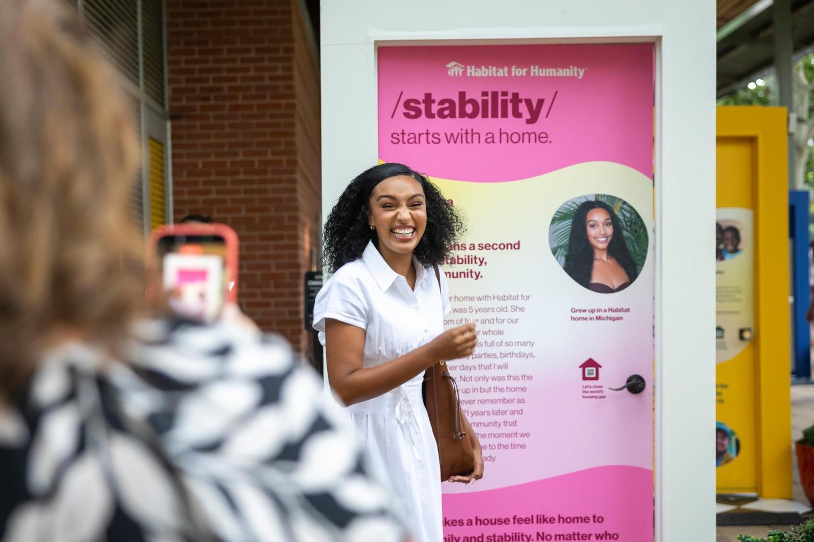 Young woman in a white dress posing in front of a pink door while someone takes a photo on their phone off frame.