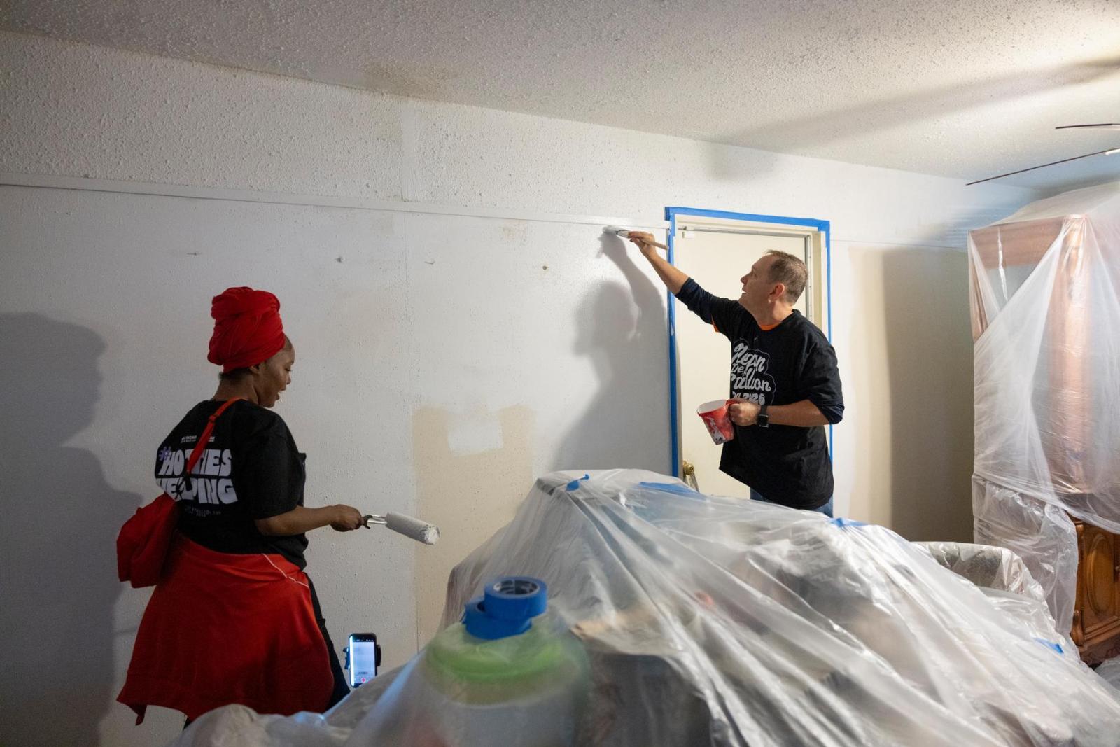 Two volunteers in black shirts walk around furniture that is wrapped in plastic to begin painting a homeowner's wall.
