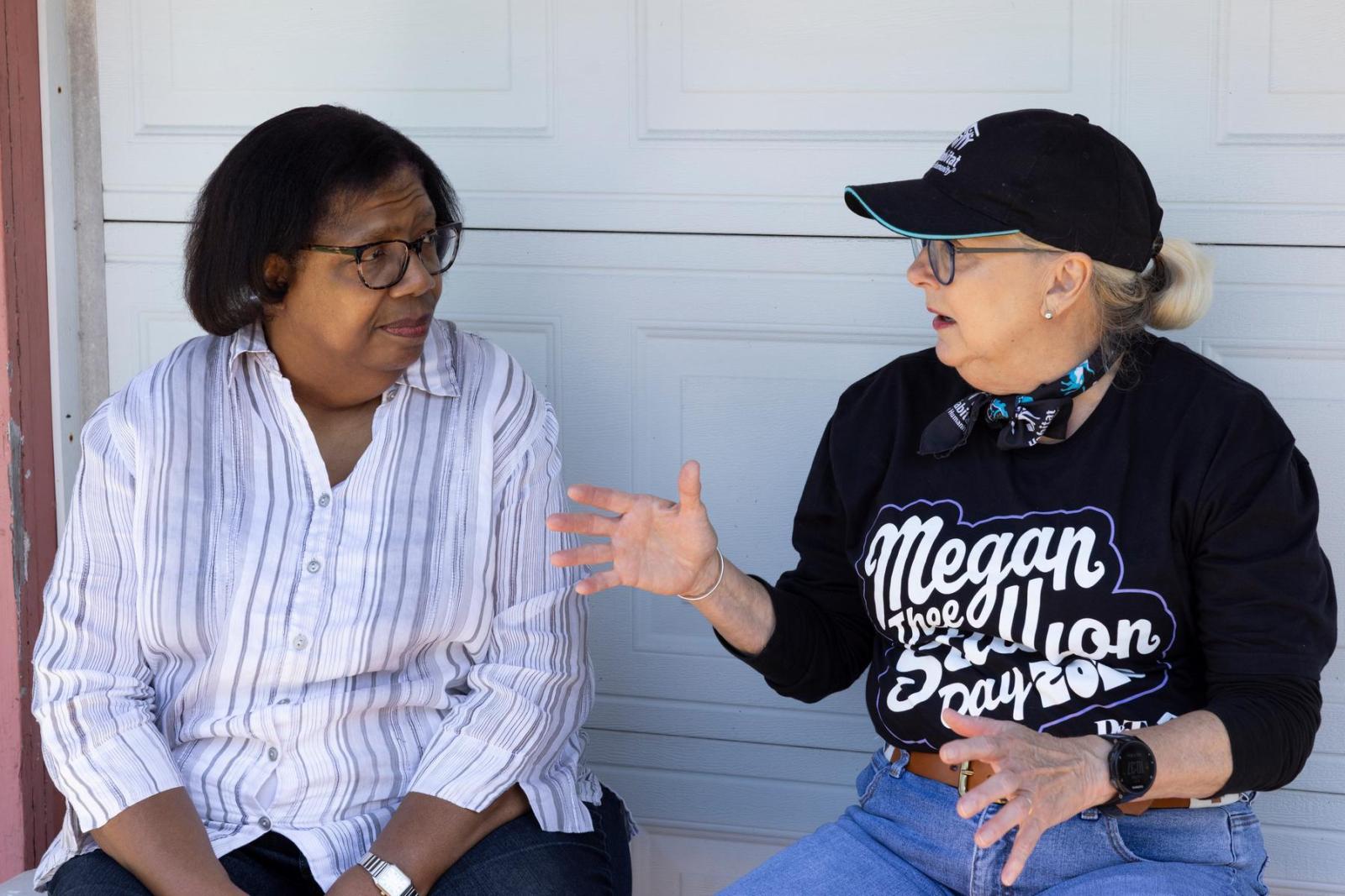 A volunteer in a black shirt and hat sits next to a homeowner and has an animated conversation. 