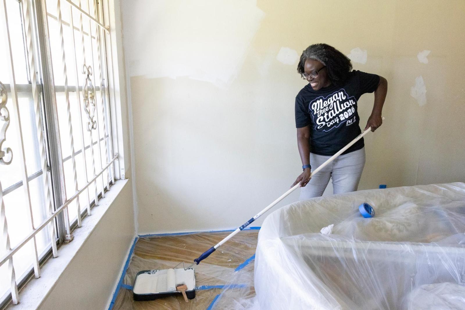 A volunteer in a black shirt stands in front of a half-painted wall and dips a long-stick paint brush into a pan.