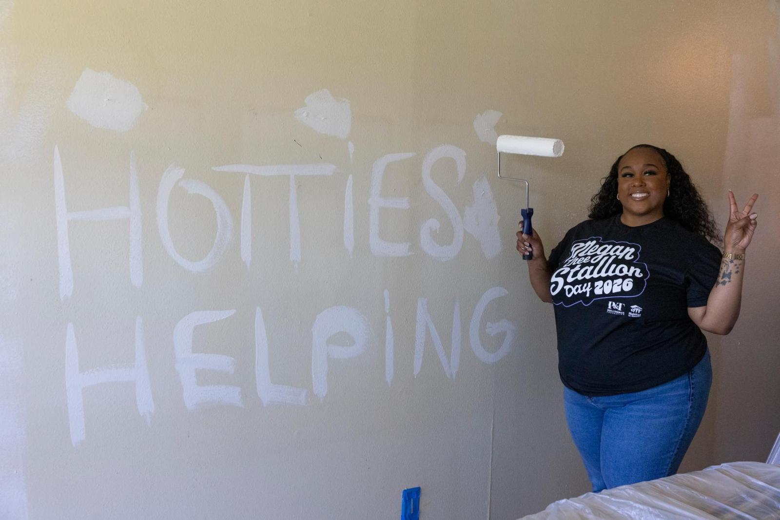 A volunteer in a black t-shirt playfully paints the phrase Hotties Helping in white before fully painting the homeowner's wall in a fresh coat of white paint.