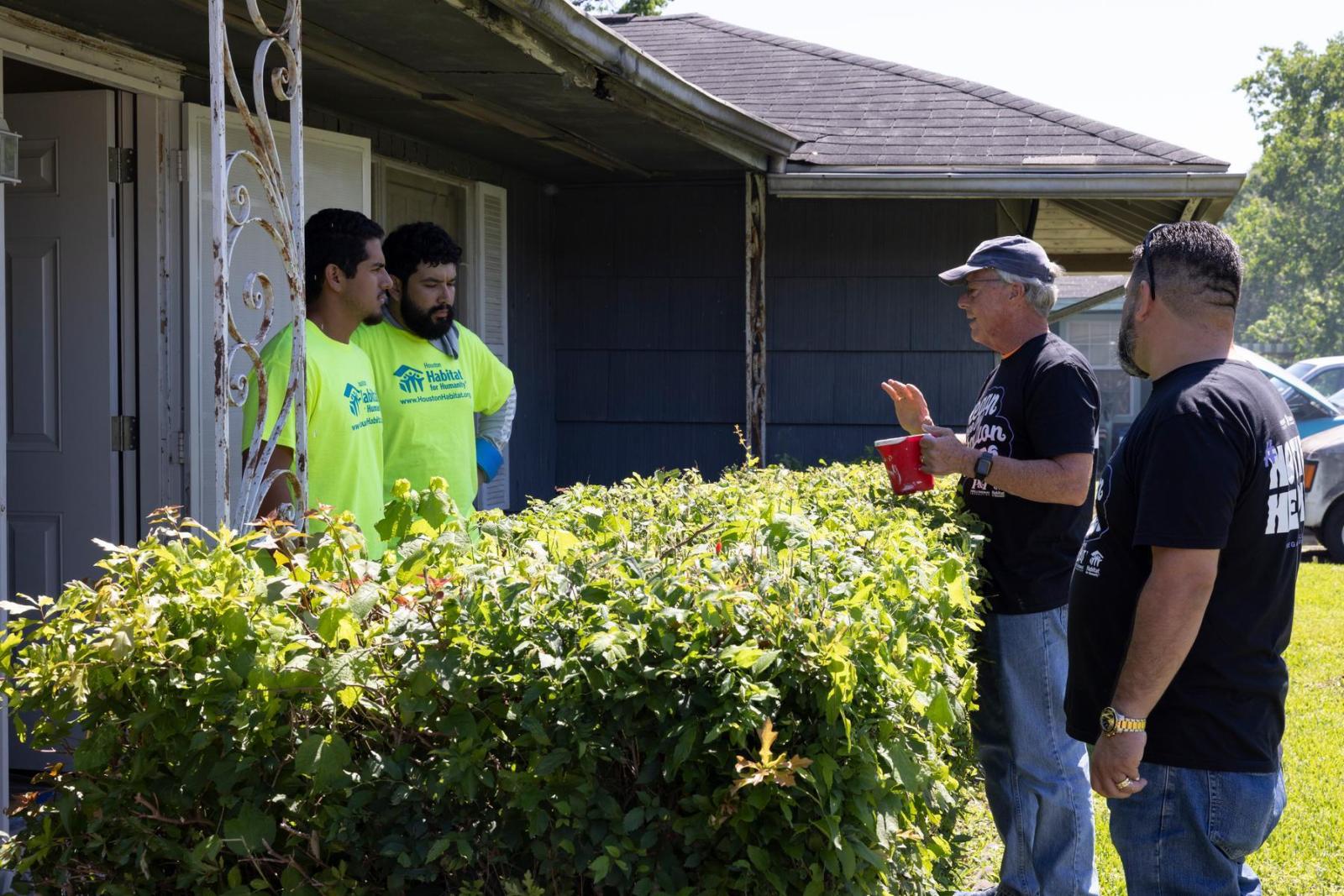 Two people in bright yellow shirts speak across a bush to a pair of volunteers in black shirts.