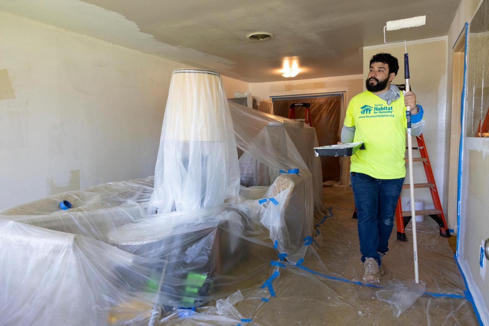 A volunteer in bright yellow Houston Habitat shirt carries a long pole and paint brush past furniture wrapped in plastic.