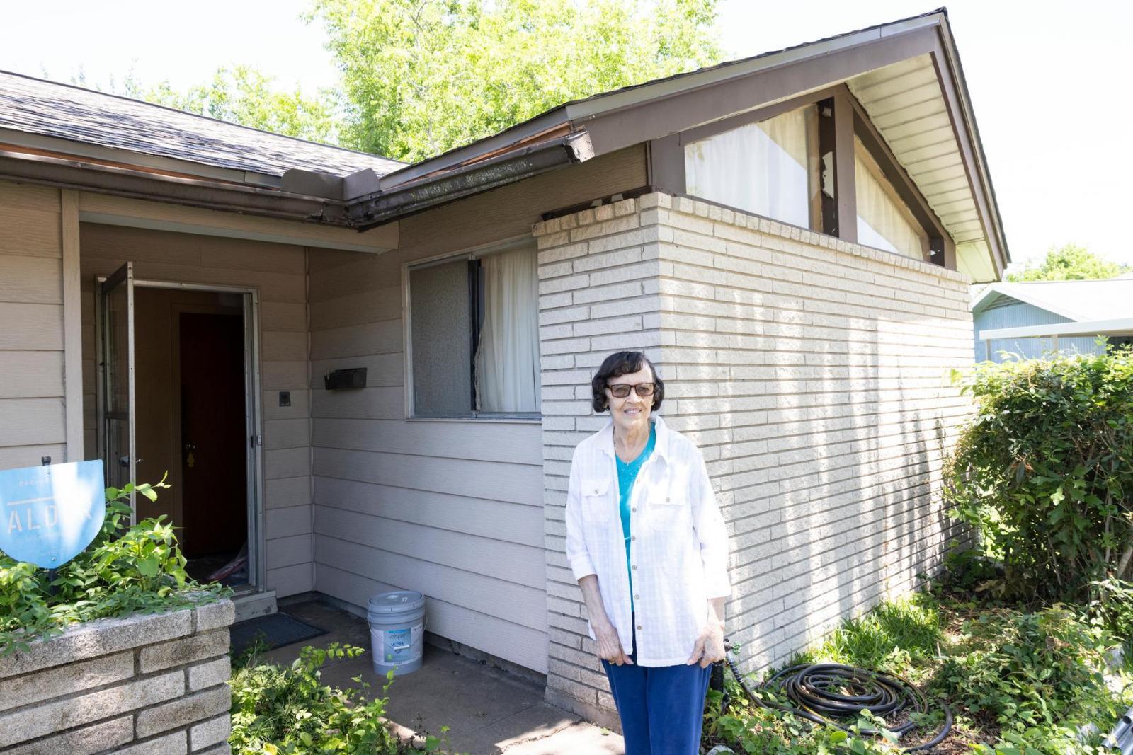 A homeowner wearing a white button down shirt stands in front of her white brick home and smiles.
