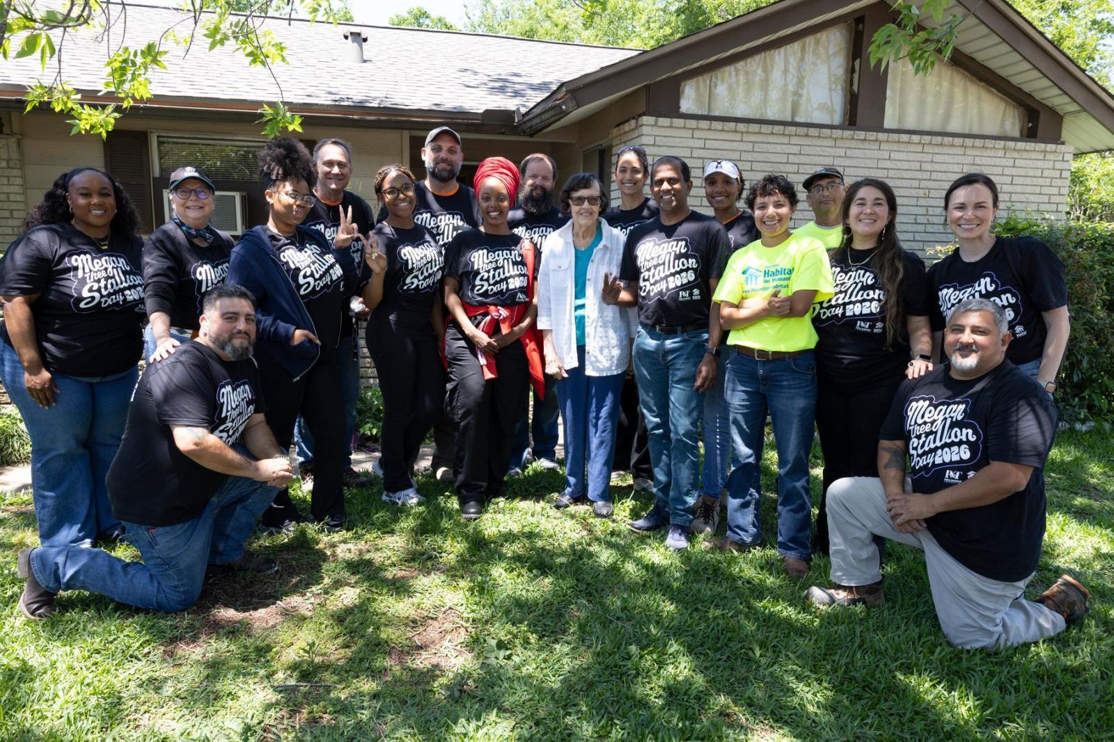 A homeowner stands in the middle of a group of volunteers wearing black and bright yellow t-shirts in front of her home.