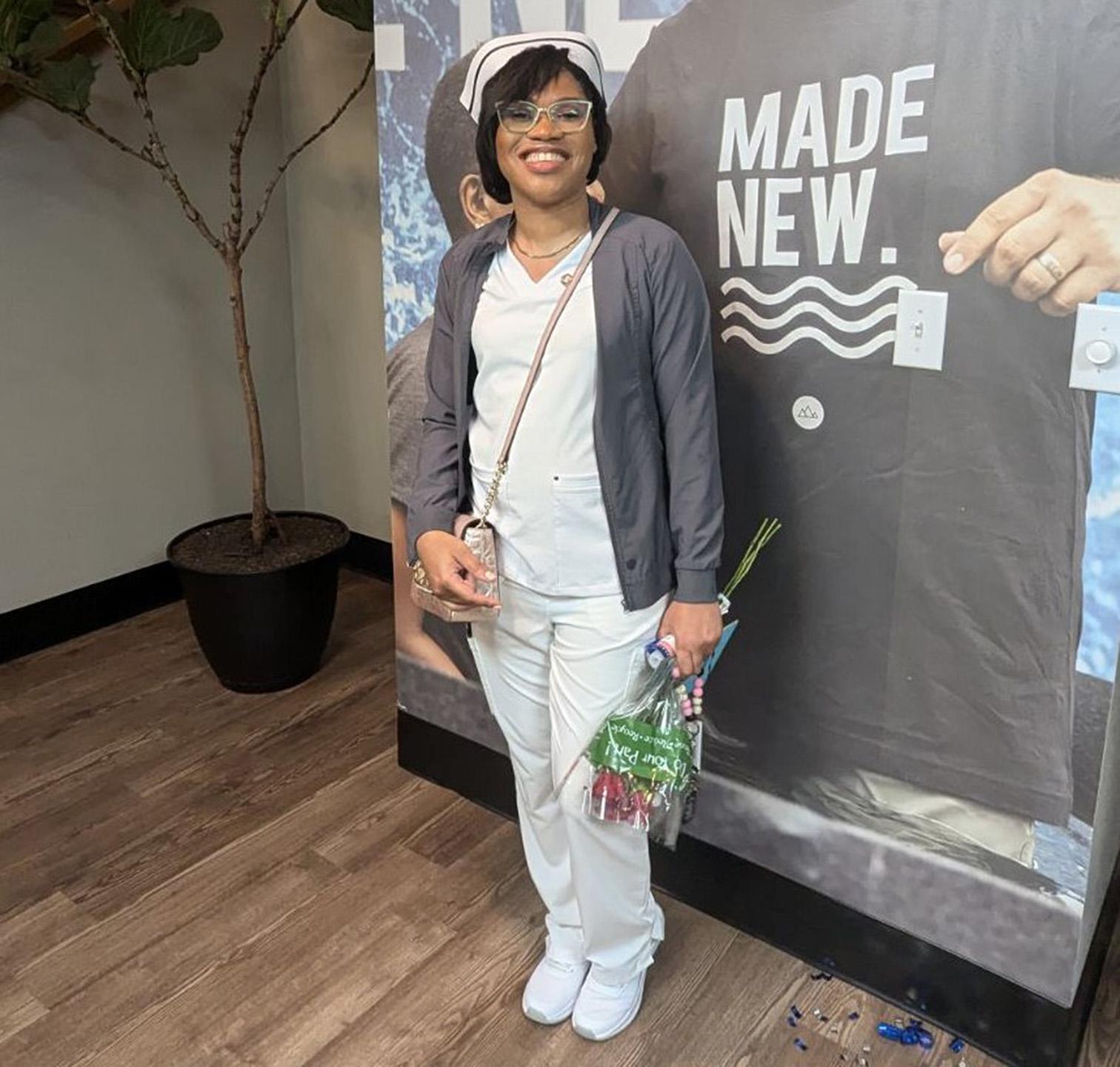 Woman in white scrubs and a gray cardigan smiling holding flowers. 