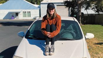 Young woman in black hat, orange sweatshirt and jeans sitting on the hood of a white car.