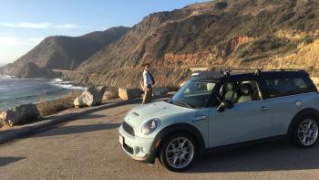 Light blue mini-cooper near a seascape and mountains with a man standing in the background.