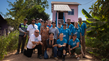 A group of Global Village volunteers wearing matching bright blue Habitat shirts pose together in front of a newly-constructed pink house and palm trees.
