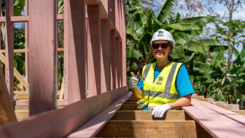 A woman in bright blue T-shirt, flourescent yellow safety vest, a white hardhat and sunglasses smiles and gives a thumbs-up while standing between painted wooden slats on a Habitat Global Village build site.