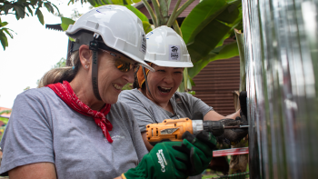 Two women volunteers in hardhats work together to screw siding onto a house