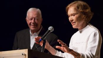 Mrs. Carter at a podium speaking into a microphone as President Carter smiles in the background.