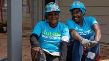 A future Habitat homeowner and a volunteer take a break on the porch of the house they're building. They are both smiling, wearing blue t-shirts and hard hats covered in stickers.