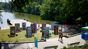 A wide shot of the art installation of pop-up doors showing door frames with art on them in a park setting with a body of water behind it. There is a sidewalk in front with people strolling.