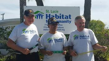 Three male veterans standing in front of a Habitat sign.  First is holding a hammer, second is holding a level, third is holding a tape measure.