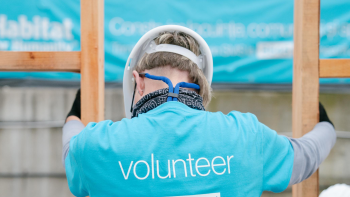 The back of a bright blue shirt that says "volunteer" is worn by a person with a hardhat and gloves while they are holding wood framing on a build site. 