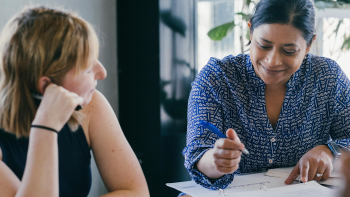 Two women sit together at a table in an office. One of the women is holding a pen while pointing at some papers on the table.