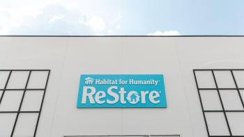 A photo of a white building and in the center is a blue sign that reads Habitat for Humanity ReStore and you can see blue skies and clouds above it.