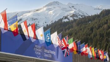 Foreground: Flags from many countries displayed over a banner with the World Economic Forum logo and text reading "Annual Meeting Davos 2026". In the background the Swiss Alps are visible, covered in snow.
