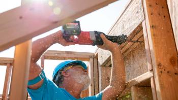 A volunteer stands with his arms above his head holding a drill that he is pushing into the frame of a house.