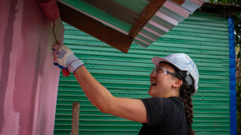 A young woman wearing a hardhat has a big smile on her face as she rolls pink paint on the side of a house.