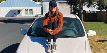 Girl in black ball cap, orange sweatshirt, leggings and tennis shoes sitting on the hood of a white car.