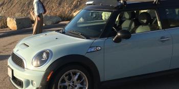 Light blue mini-cooper near a seascape and mountains with a man standing in the background.