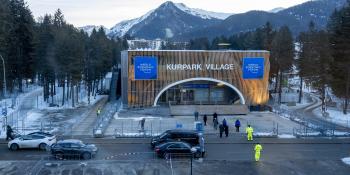 High angle photo showing the outside of the World Economic Forum venue surrounded by trees, with the Swiss Alps mountains visible in the background. The building sign says "Kurpark Village" in white text, and a blue banner is hung on either side with the World Economic Forum logo.