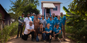 A group of Global Village volunteers wearing matching bright blue Habitat shirts pose together in front of a newly-constructed pink house and palm trees.